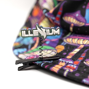May include: A black metal alligator clip attached to a fabric hat with a colorful mushroom pattern. A thin metal spring extends upward from the clip, supporting a white and grey emblem featuring the text ILLENIUM in a stylized font.