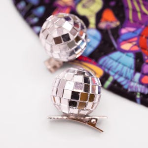 May include: Two silver disco ball hair clips. Each clip features a small, spherical disco ball attached to a metal clip. The background includes a colorful fabric with a psychedelic mushroom pattern.
