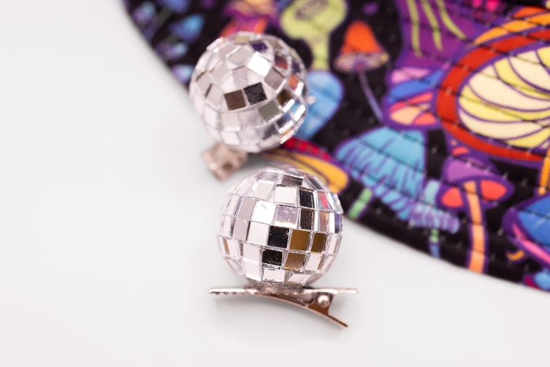 May include: Two silver disco ball hair clips. Each clip features a small, spherical disco ball attached to a metal clip. The background includes a colorful fabric with a psychedelic mushroom pattern.