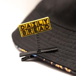 May include: A black bucket hat with a yellow sign that reads "DAILY BREAD" attached with a spring and clip. The hat has a patterned trim.