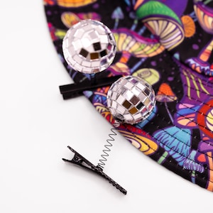 May include: A black hair clip with a silver spring and two disco ball hair accessories. The clip is next to a black fabric with a colorful mushroom pattern in purple, orange, and blue.