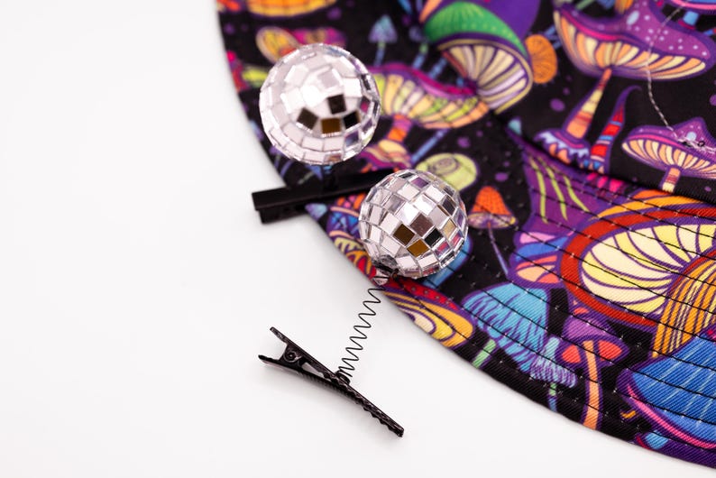 May include: A black hair clip with a silver spring and two disco ball hair accessories. The clip is next to a black fabric with a colorful mushroom pattern in purple, orange, and blue.