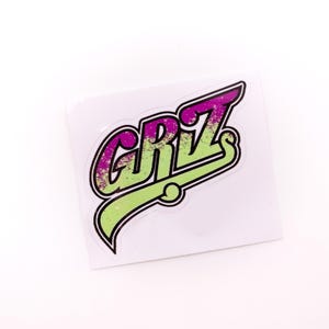 Puede incluir: Una pegatina con la palabra "GRiZ" en una fuente estilizada. Las letras están delineadas en negro, con una combinación de colores verde y morado. La pegatina está sobre un fondo blanco.