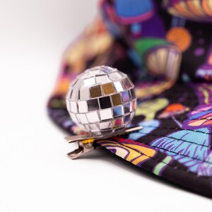 May include: A silver disco ball clip-on accessory. The clip is attached to a black hat with a colorful mushroom and psychedelic design. The hat features shades of purple, orange, and blue.