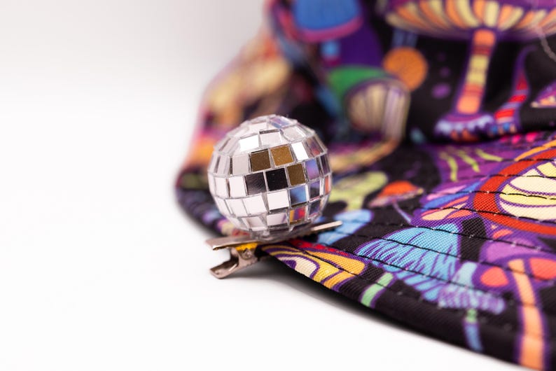 May include: A silver disco ball clip-on accessory. The clip is attached to a black hat with a colorful mushroom and psychedelic design. The hat features shades of purple, orange, and blue.