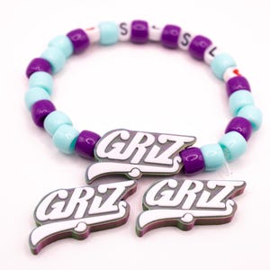 Puede incluir: Una pulsera de cuentas azules claras, moradas y blancas. La pulsera tiene la palabra "SOL" deletreada con cuentas de letras blancas. También se incluyen tres colgantes con el logotipo "GRIZ" a juego.