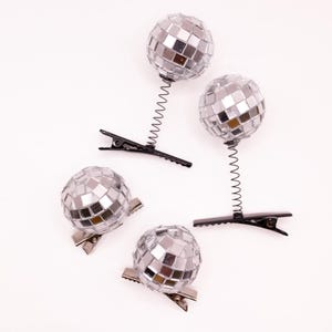 May include: Four silver disco ball hair clips. Two clips have a black spring and clip base, while the other two have a silver clip base. The disco balls are made of small, reflective mirrored squares.
