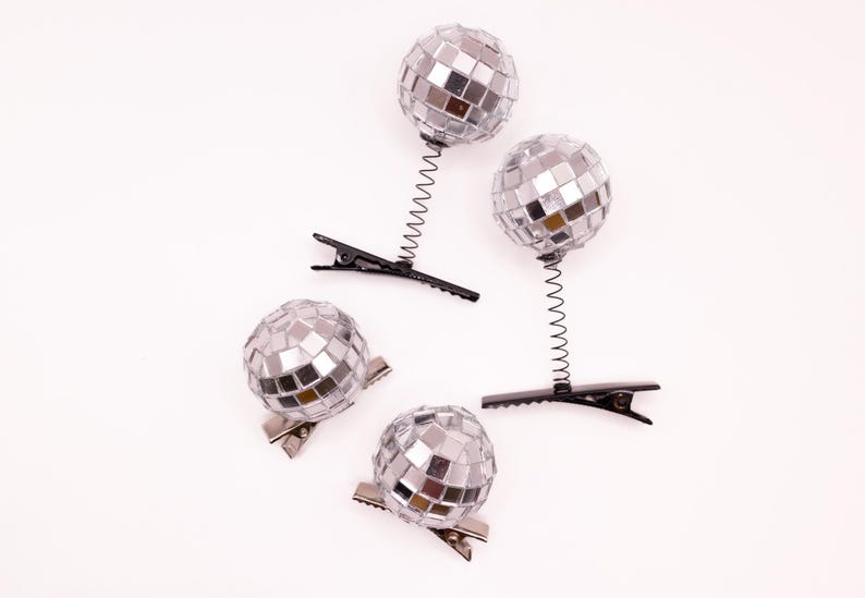 May include: Four silver disco ball hair clips. Two clips have a black spring and clip base, while the other two have a silver clip base. The disco balls are made of small, reflective mirrored squares.