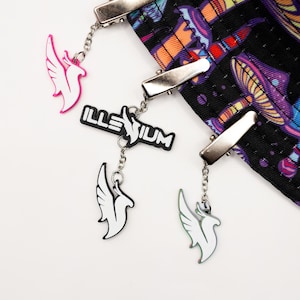May include: Three silver metal clips attached to a black fabric with a colorful mushroom pattern. Each clip features a dangling charm: a pink phoenix, a black and white Illenium logo with a white phoenix, and a white phoenix charm.