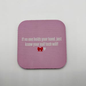 May include: A pink square coaster with rounded corners. The coaster features the text "If no one holds your hand, just know your nail tech will!" in white, with a red bow and a white heart.