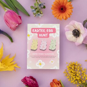Pode incluir: Um cartão rosa com o texto "Easter Egg Hunt" e três ilustrações de coelhos de desenho animado. O cartão tem um design floral rosa e branco. O cartão é cercado por várias flores, incluindo tulipas rosa, mimosa amarela e anémones brancas.