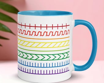 Puede incluir: Taza de cerámica blanca con interior y asa azules. La taza presenta un diseño geométrico colorido con bandas horizontales rojas, naranjas, amarillas, verdes y moradas. El diseño recuerda a un arcoíris.