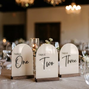 May include: Three table number signs with a cream-colored arched top and a dark wooden base. The words "Table One", "Table Two", and "Table Three" are written in black script. The signs are set on a table with glasses and flowers.