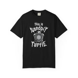 May include: Black t-shirt with a white graphic design. The design features the text "This is Sandpit Turtle" in a stylized font, along with an illustration of a turtle with a circular pattern on its shell. The shirt has short sleeves and a crew neck.