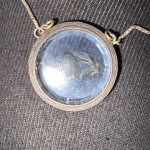 Puede incluir: Un colgante redondo de plata con fondo azul y diseño floral. El colgante tiene una cubierta de vidrio transparente y está unido a una cadena de plata. El marco del colgante tiene un diseño ornamentado y grabado.