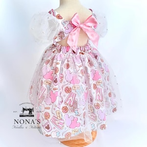 May include: A white tulle dress with pink and brown Mickey Mouse print. The dress has a pink satin bow on the back and pearl embellishments on the tulle.