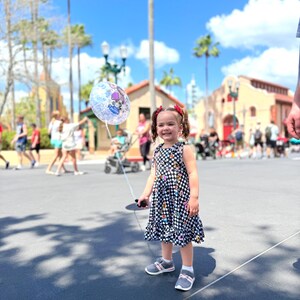 May include: A young girl in a black and white checkered dress with colorful flowers and a cartoon character print holds a clear balloon with a cartoon character on it. She is smiling and looking at the camera. The girl is standing on a paved walkway in front of a building with a red roof.