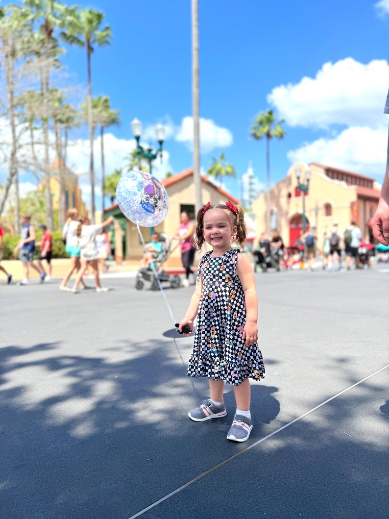 May include: A young girl in a black and white checkered dress with colorful flowers and a cartoon character print holds a clear balloon with a cartoon character on it. She is smiling and looking at the camera. The girl is standing on a paved walkway in front of a building with a red roof.