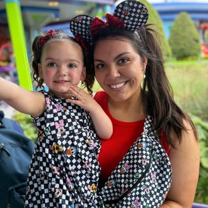 May include: A woman and a young girl wearing matching black and white checkered dresses with Disney character prints. The woman is wearing a red tank top and a black and white checkered fanny pack with Disney character prints. Both are wearing black and white checkered mouse ears with red bows.