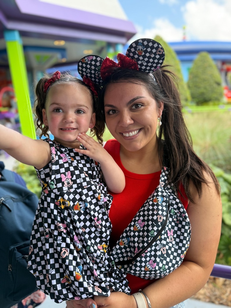 May include: A woman and a young girl wearing matching black and white checkered dresses with Disney character prints. The woman is wearing a red tank top and a black and white checkered fanny pack with Disney character prints. Both are wearing black and white checkered mouse ears with red bows.