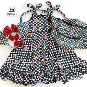 May include: A black and white checkered dress with a cartoon character print. The dress has a halter neck style and a ruffled hem. The dress is paired with a matching black and white checkered fanny pack and a headband with a red bow.