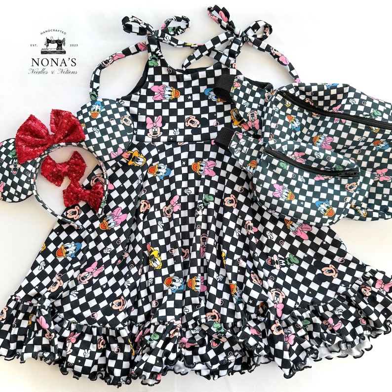 May include: A black and white checkered dress with a cartoon character print. The dress has a halter neck style and a ruffled hem. The dress is paired with a matching black and white checkered fanny pack and a headband with a red bow.