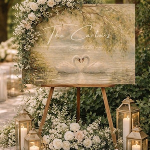 May include: A wooden easel displays a sign with the text "The Carters Est 2020" above a painting of two swans. The sign is decorated with white roses and greenery. Gold lanterns with candles are placed around the easel.