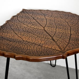 May include: A wooden table with a leaf-shaped top and black metal hairpin legs. The table top is intricately carved with a leaf vein pattern.