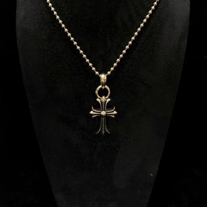 May include: Silver-toned cross pendant necklace. The pendant features a detailed cross design with ornate embellishments. The necklace has a ball chain. The cross is suspended from a circular bail. The background is black.