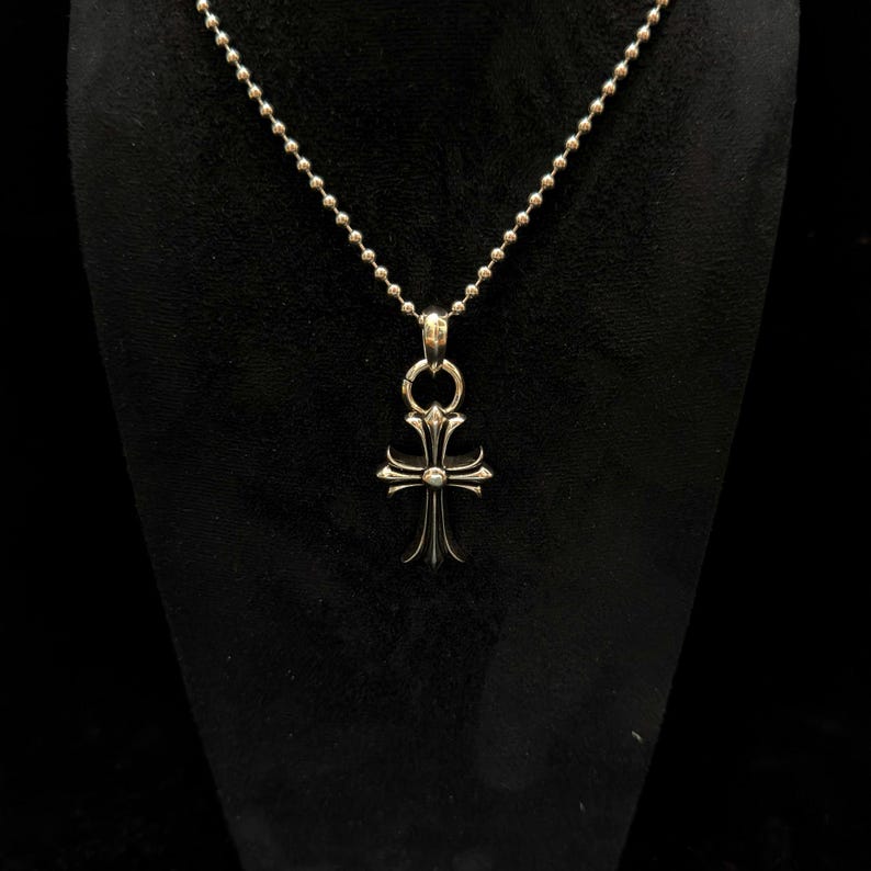 May include: Silver-toned cross pendant necklace. The pendant features a detailed cross design with ornate embellishments. The necklace has a ball chain. The cross is suspended from a circular bail. The background is black.