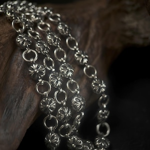 May include: Close-up of a silver necklace with a floral design. The necklace is composed of multiple strands of small, flower-shaped links connected by circular rings. The metal has a polished, reflective surface, and the necklace is displayed against a dark, textured background.