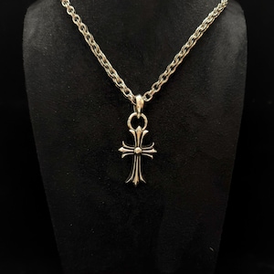 May include: Silver-toned necklace with a cross pendant. The pendant features a detailed cross design with pointed ends and a central embellishment. The chain is a textured, linked style. The necklace is displayed against a dark background.