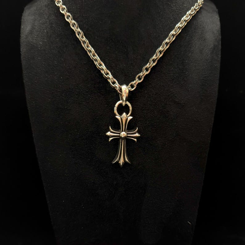 May include: Silver-toned necklace with a cross pendant. The pendant features a detailed cross design with pointed ends and a central embellishment. The chain is a textured, linked style. The necklace is displayed against a dark background.