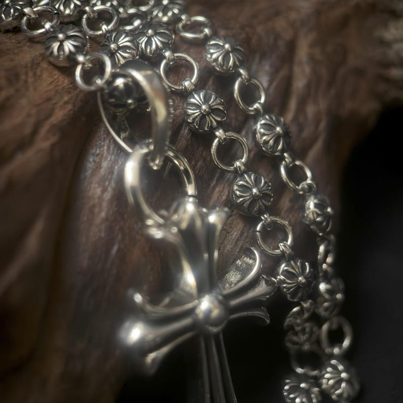 May include: Close-up of a silver necklace with a cross pendant and floral-shaped links. The necklace is made of a shiny metal and is displayed against a dark, textured background. The cross pendant is detailed and ornate.
