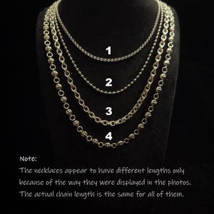 May include: Four silver necklaces displayed on a black stand. The necklaces have different chain styles, including a ball chain and a chain with decorative elements. The image includes text that explains the necklaces appear to have different lengths only because of the way they were displayed.
