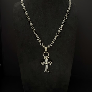 May include: A silver necklace with a cross pendant. The chain is made of small, round beads. The cross pendant has a detailed design with curved edges and a central element. The necklace is displayed against a black background.