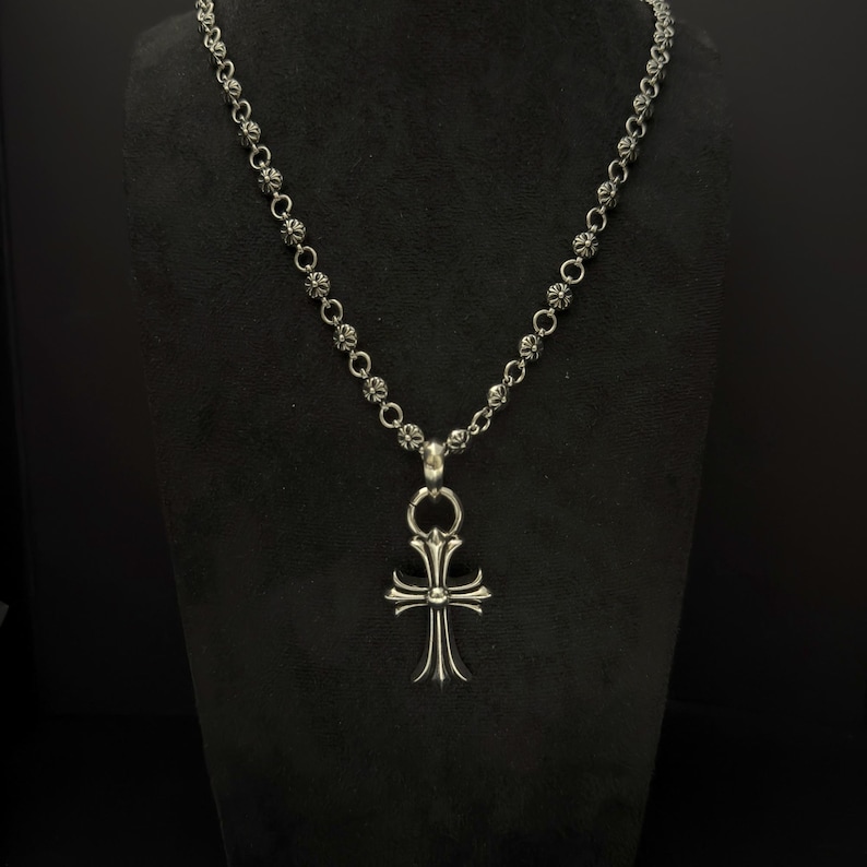 May include: A silver-colored necklace with a cross pendant. The necklace features a chain of small, round beads. The cross pendant has a detailed design with curved edges and a central element. The necklace is displayed against a black background.