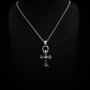 May include: A silver-colored pendant necklace with a cross-shaped charm. The charm has a detailed design with a central sphere and ornate, pointed ends. The chain is a delicate, linked style, and the background is black.