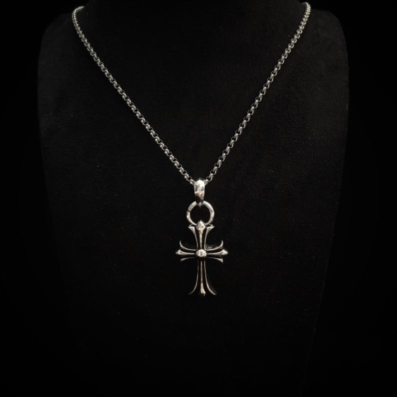 May include: A silver-colored pendant necklace with a cross-shaped charm. The charm has a detailed design with a central sphere and ornate, pointed ends. The chain is a delicate, linked style, and the background is black.