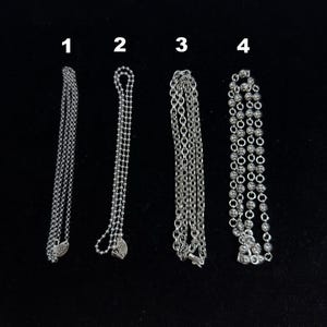 May include: Four silver necklaces displayed against a black background. The necklaces vary in design, including a delicate chain, a beaded chain, a classic link chain, and a chain with decorative ball accents. Each necklace is a different style.