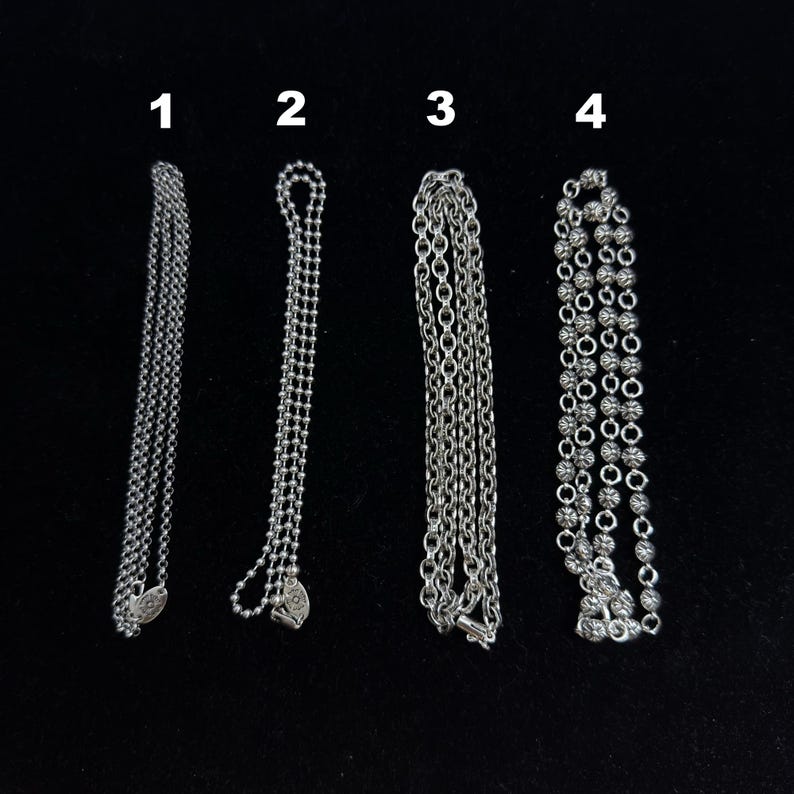 May include: Four silver necklaces displayed against a black background. The necklaces vary in design, including a delicate chain, a beaded chain, a classic link chain, and a chain with decorative ball accents. Each necklace is a different style.