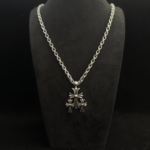 May include: Silver necklace featuring a triple cross pendant. The chain is a linked design, and the pendant has three ornate crosses. The necklace is displayed on a black mannequin.