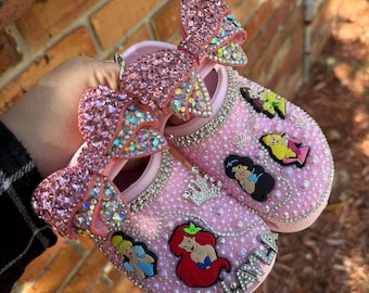 Puede incluir: Zapatos de bebé rosas adornados con brillantes pedrería, aplicaciones de personajes de princesas y grandes lazos a juego. Los zapatos presentan un ribete de perlas y un diseño de tiara. Un diseño encantador y caprichoso.