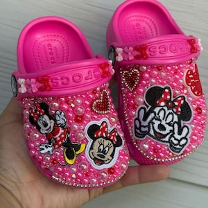 May include: Bright pink Crocs clogs, decorated with pink and clear beads. The clogs feature Minnie Mouse patches and red heart accents. Floral and heart-shaped charms are attached to the straps.