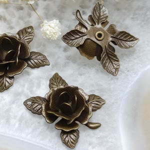 Può includere: Primo piano di tre elementi decorativi in metallo color bronzo antico a forma di rosa. Ogni rosa ha petali dettagliati e accenti di foglie. Un elemento ha un foro centrale, gli altri sono a forma di rosa.