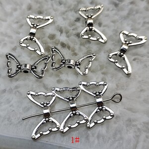 Può includere: Componenti di gioielleria in metallo color argento a forma di farfalla. I componenti hanno un design traforato dettagliato con bordi arrotondati e piccole perline decorative. L'immagine include diversi pezzi singoli e un gruppo infilato su un filo. Il numero "1#" è visibile.