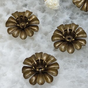 Può includere: Diversi componenti a forma di fiore color ottone antico. Ogni componente presenta un design dettagliato dei petali e una struttura centrale con molteplici piccole sporgenze. I componenti sono disposti su una superficie marmorizzata bianca e grigia.