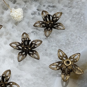 Può includere: Primo piano di diversi componenti a forma di fiore in filigrana color bronzo. Ogni componente ha cinque petali con intricati disegni ritagliati. I componenti sono sparsi su una superficie chiara e marmorizzata.