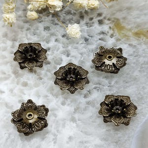 Può includere: Cinque copri perline a forma di fiore color bronzo antico. Ogni cappuccio presenta un design floreale dettagliato con un foro centrale per l'infilatura. I cappucci sono disposti su una superficie bianca e grigia con fiori secchi.