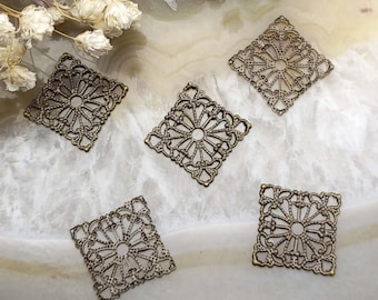 May include: Five bronze-colored filigree square charms with an openwork floral design. The charms are arranged on a light-colored, marbled surface, with dried flowers in the background. These decorative metal charms are suitable for jewelry making and crafting.